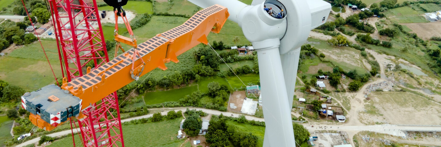 Ein Krahn steht vor einer Windenergieanlage. An dieser fehlt einer von drei Rotoren. Aus der Verbindungsstelle schauen drei Monteur*innen heraus.