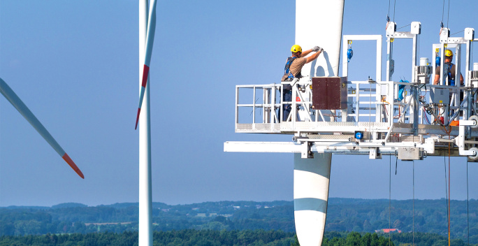 Ein Windrad und im Vordergrund arbeitet jemand auf einer Plattform an einem Rotor in luftiger Höhe