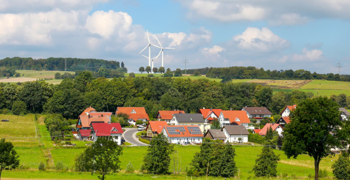 Vor mehreren Windrädern liegt ein Dorf, auf dessen Dächer Solarmodule angebracht sind.