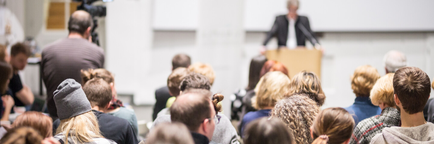 Das Bild zeigt einen Vortrag in einem Saal, bei dem ein Redner vorne an einem Pult spricht. Das Publikum sitzt dicht beisammen und hört aufmerksam zu, während eine Kamera die Szene aufzeichnet.