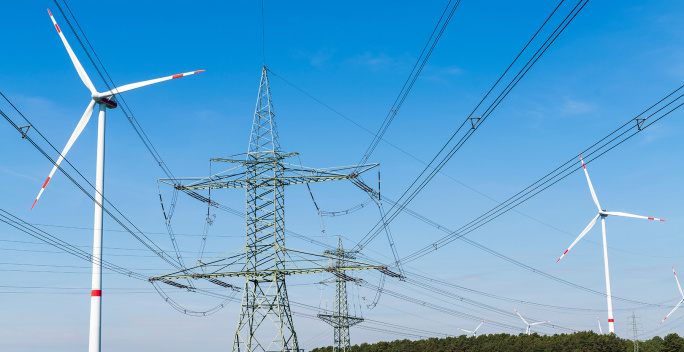 Auf einer Wiese befinden sich mehrere Windräder. Zwischen ihnen befinden sich Freileitungsmasten.