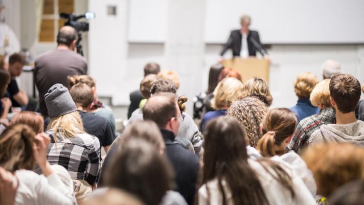 Das Bild zeigt einen Vortrag in einem Saal, bei dem ein Redner vorne an einem Pult spricht. Das Publikum sitzt dicht beisammen und hört aufmerksam zu, während eine Kamera die Szene aufzeichnet.