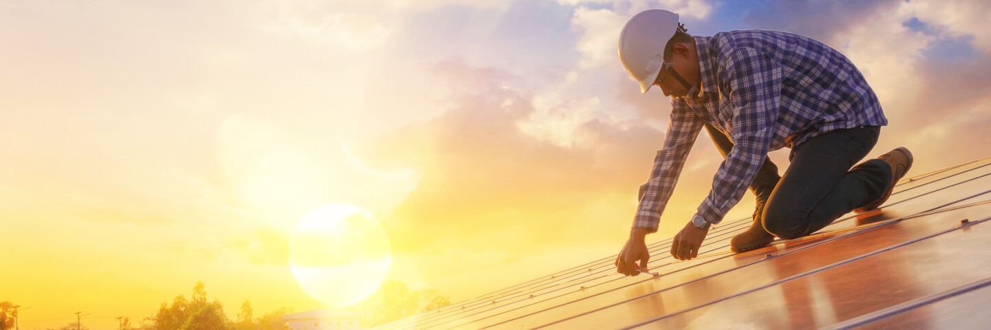 Mann kniet bei Sonnenuntergang auf einer Photovoltaikanlage und schraubt ein Panel fest.